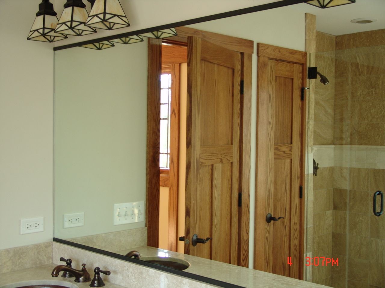 Bathroom mirror reflecting wooden doors, sink, and shower. Black trim, neutral walls, warm wood tones.