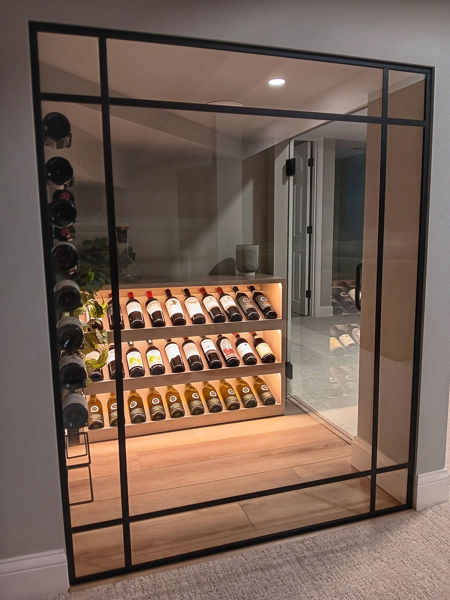 Wine cellar behind a black metal and glass frame. Rows of wine bottles on shelves and side racks, lit from within.