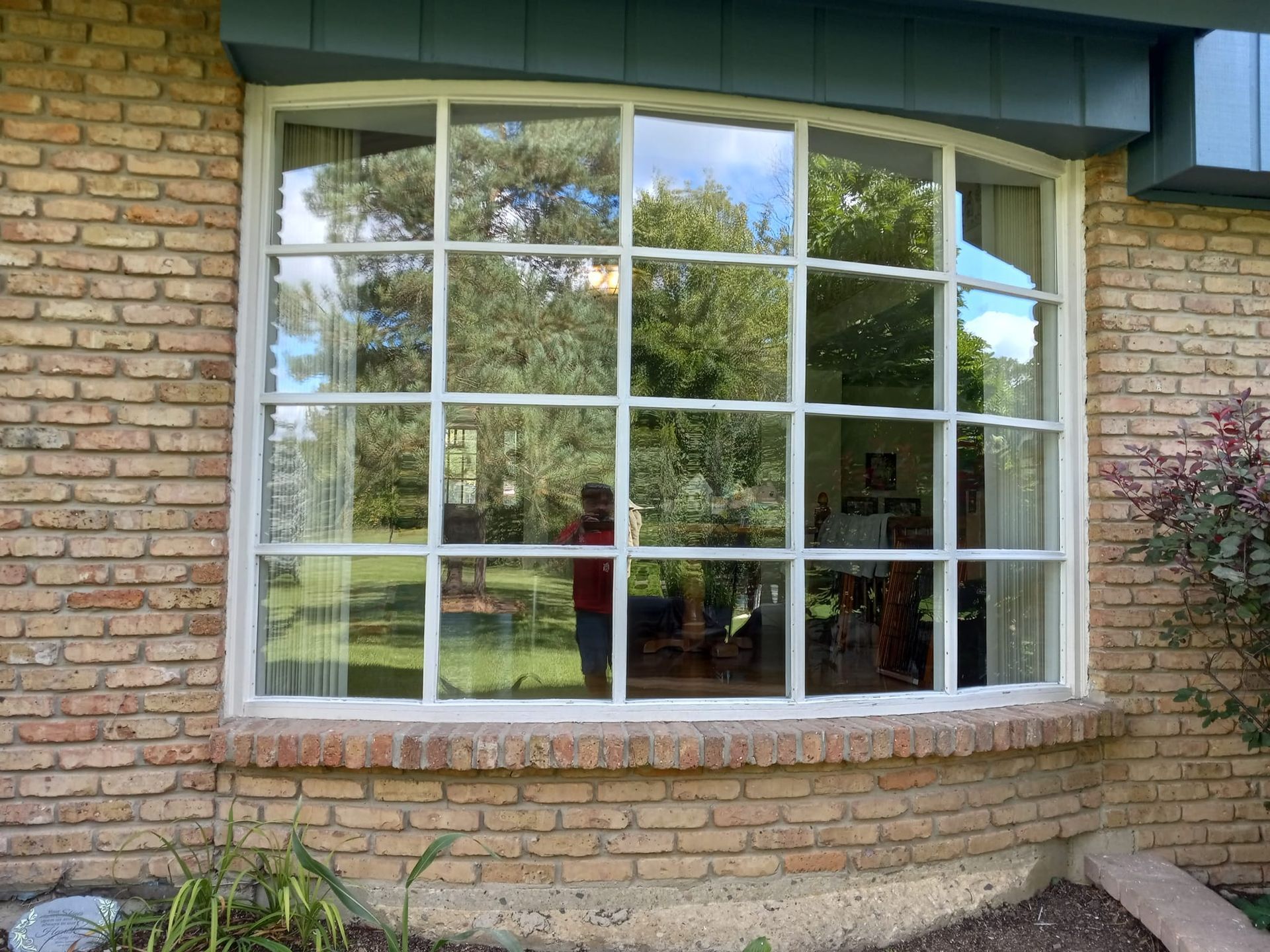 Large multi-pane window in a brick building; a person is reflected in the glass, trees outside.