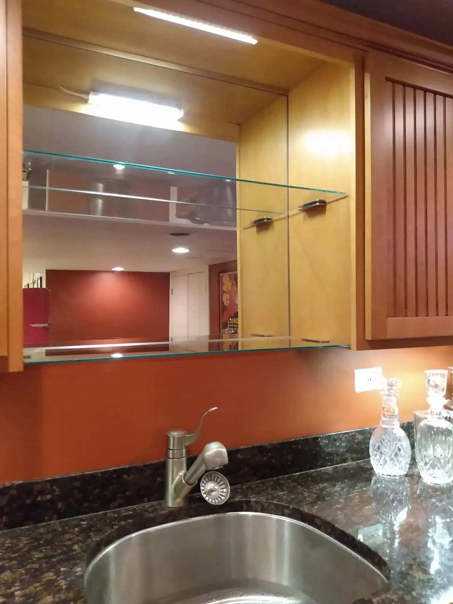 Bar sink with cabinet, glass shelves, and mirror. Orange wall and granite countertop.