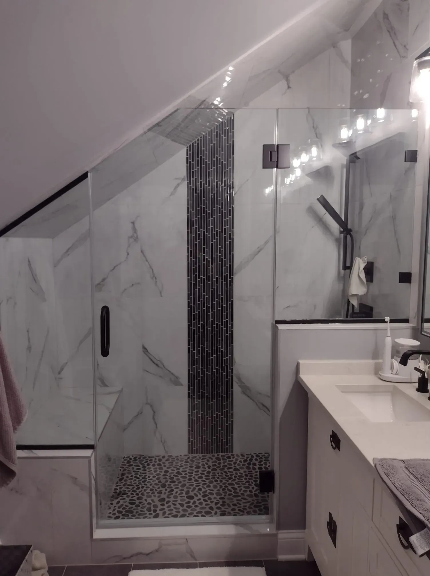 Modern bathroom with glass shower, marble tile, and pebble floor. Angled ceiling.
