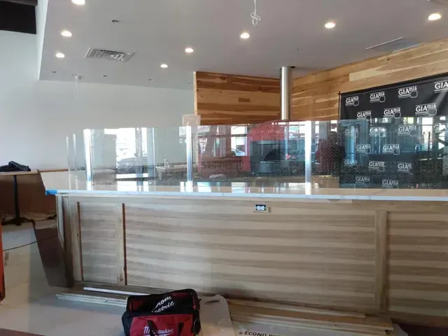 Interior view of a business counter with clear acrylic protective barriers, wooden paneling, and a light-colored countertop.