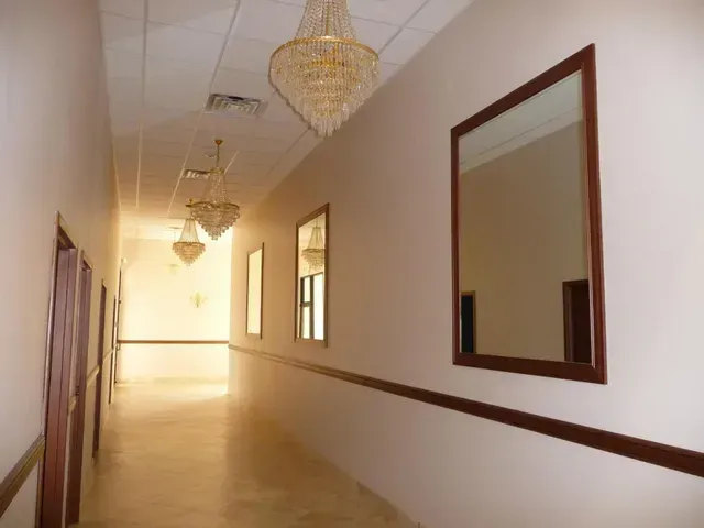 Long hallway with mirrors and chandeliers; beige walls, brown trim.