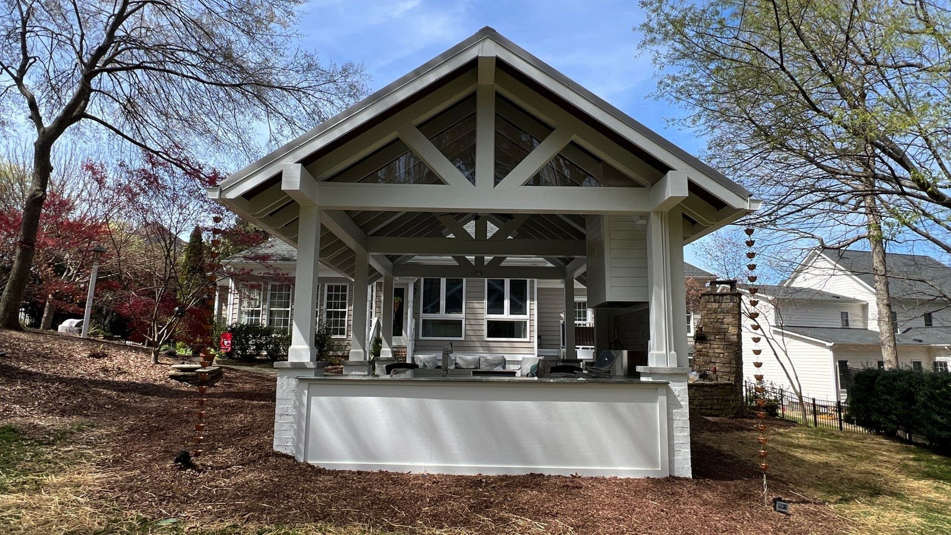 outdoor kitchen with white structure, grill, and seating under a gabled roof in a backyard
