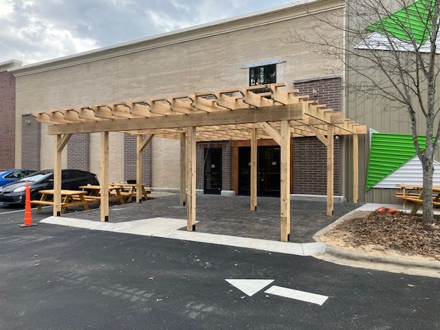 Pergola entrance to a brick building with picnic tables, on a paved area.