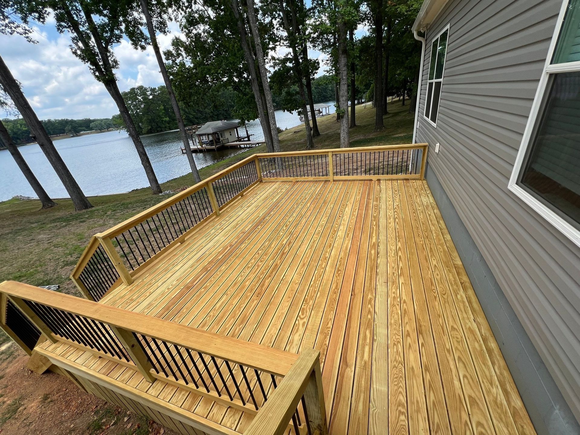 a wooden deck with black railing on the side of a gray house overlooking a lake