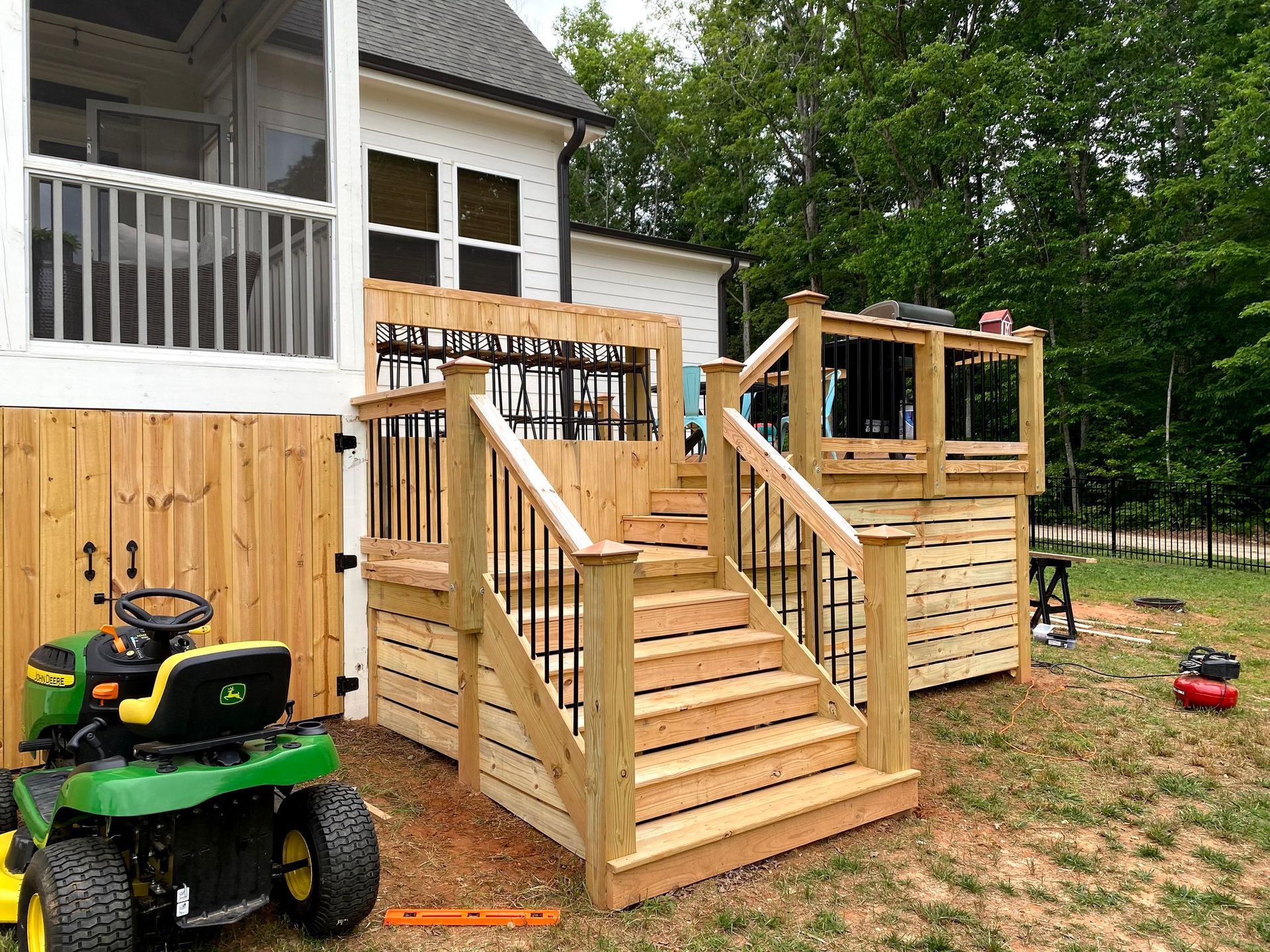 a wooden deck and staircase with black railings, beside a lawnmower, near a house