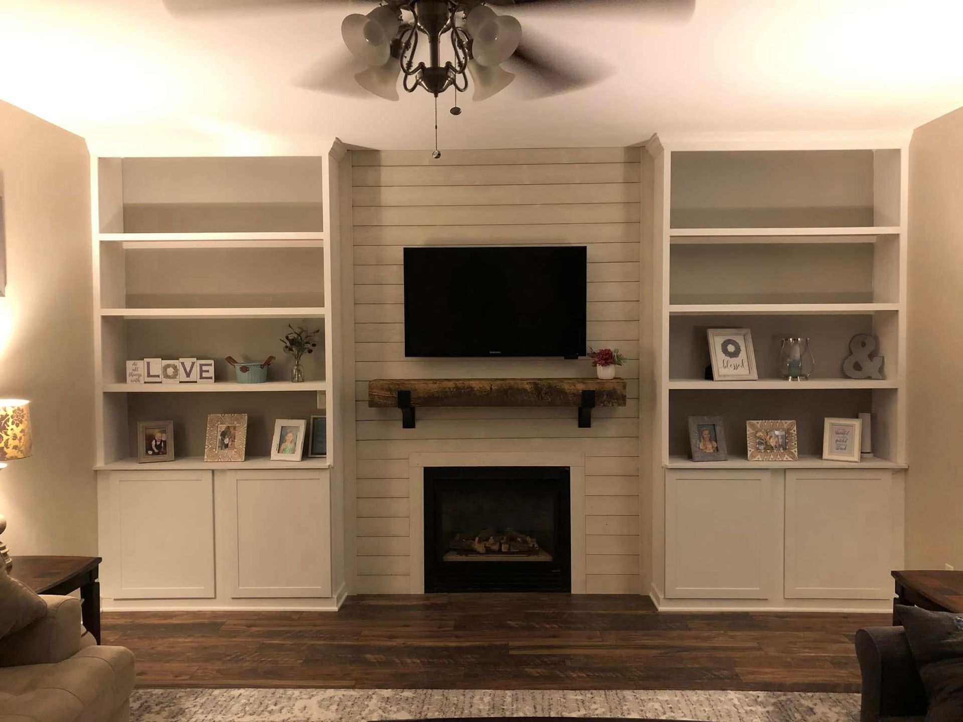 A built-in white bookshelves flank a fireplace with a TV, a rustic wooden mantel, and shiplap accent wall