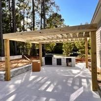 A wooden pergola over a patio with a grill and trees in the background.