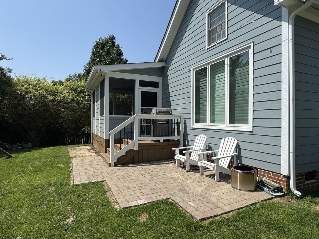 a backyard patio with a screened porch, white chairs, and a blue house on a sunny day