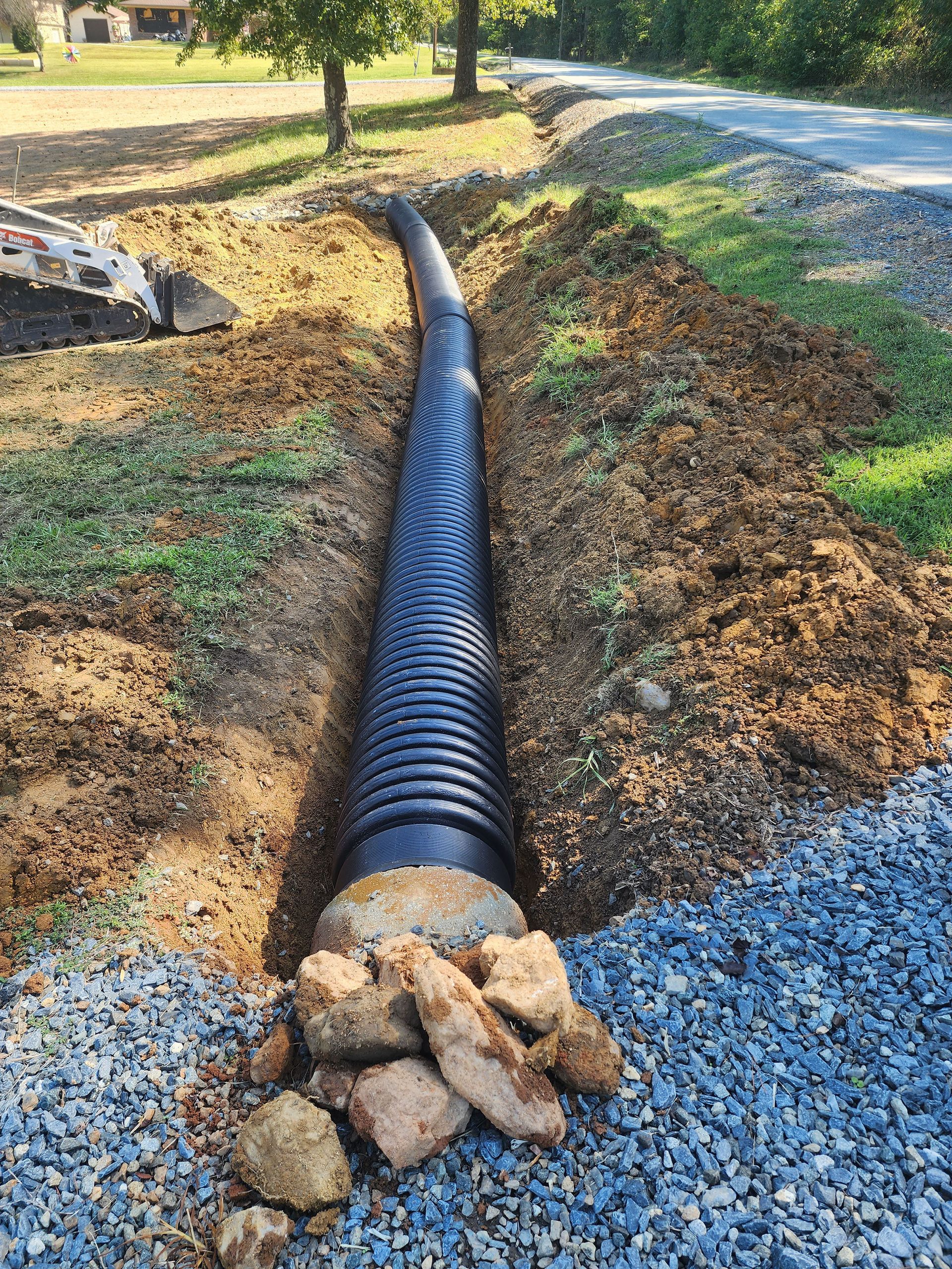 A black pipe is being installed in the dirt next to a gravel road.