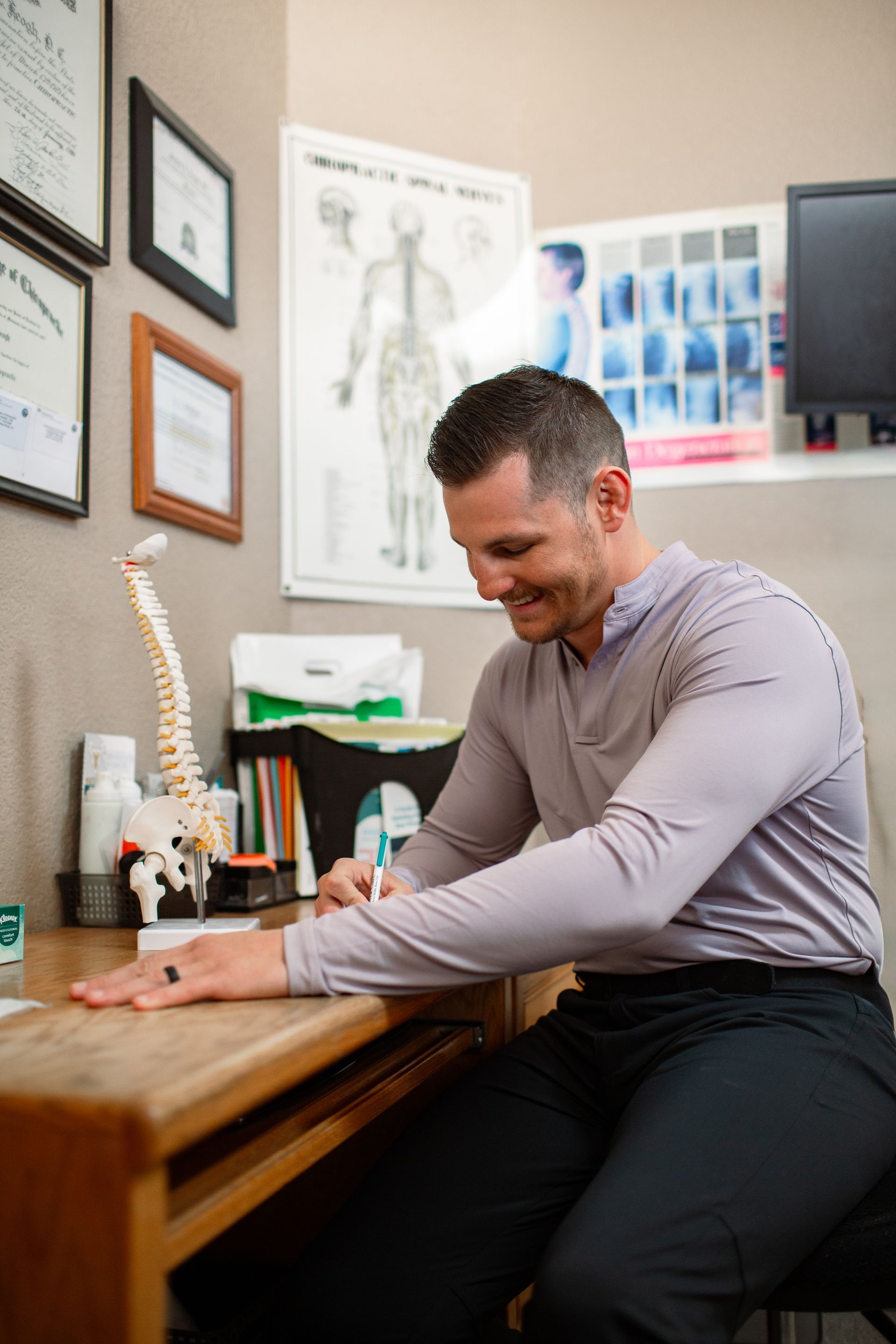 Man in gray shirt at a desk, writing. Office setting, spinal model visible. Smiling.