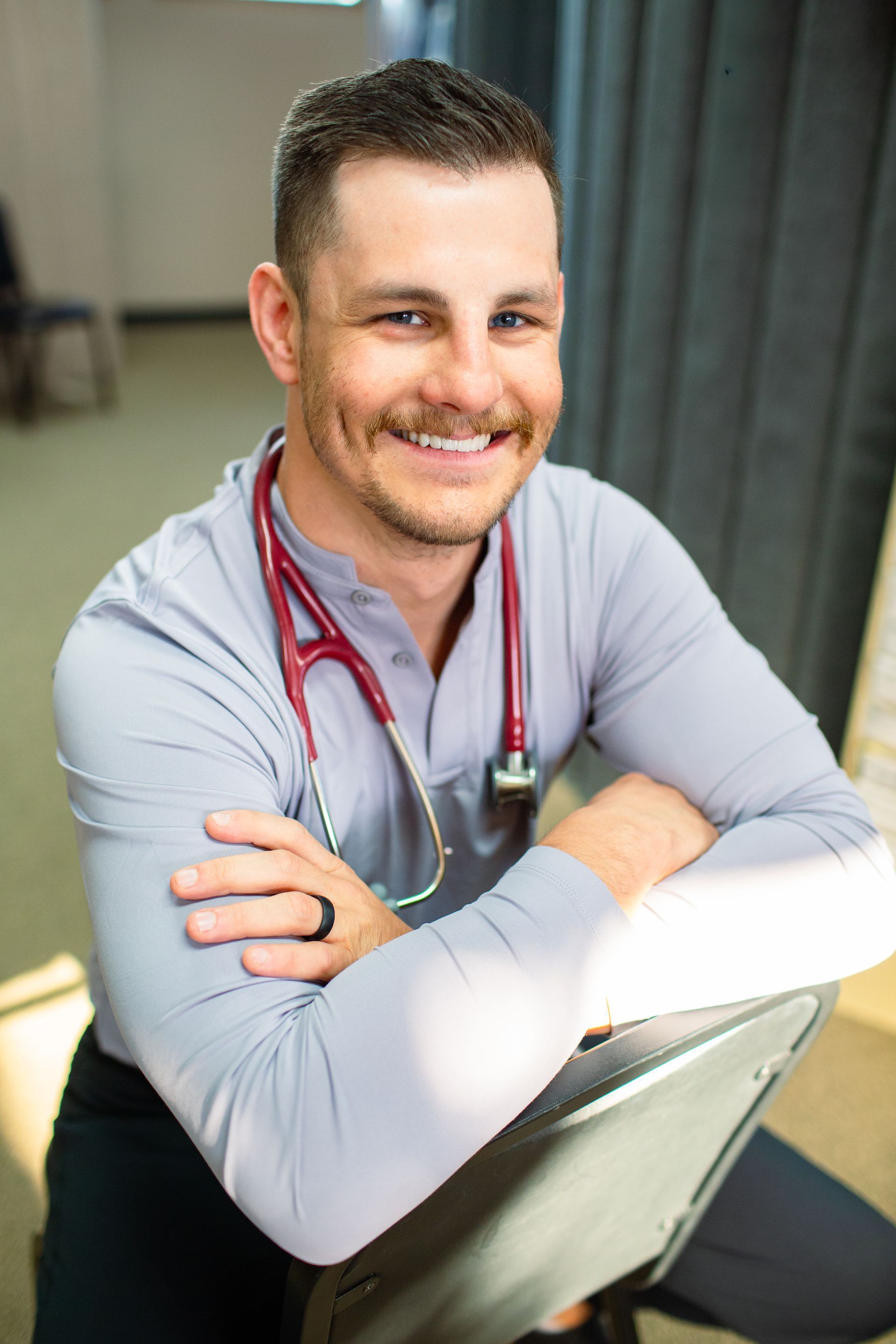 Man with stethoscope smiles, leaning back in chair. Gray shirt, red stethoscope.