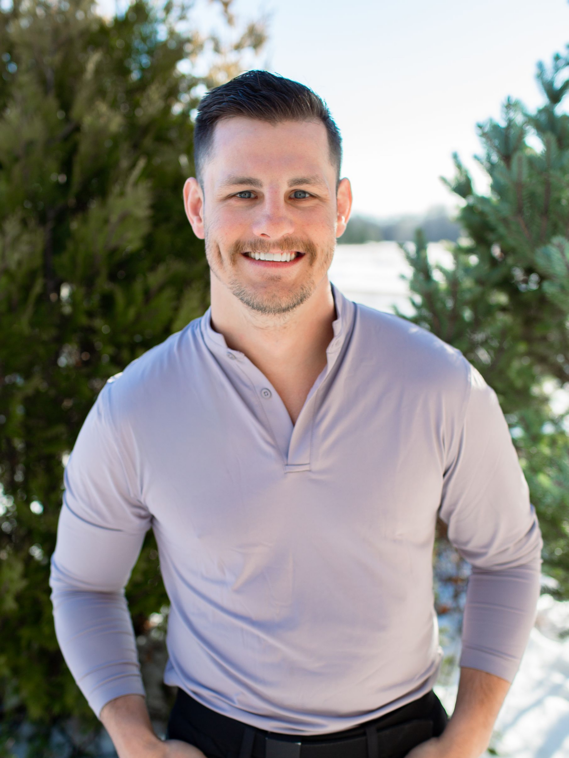 Man with hands in pockets smiling outdoors, wearing a gray shirt and black pants, snow in background.