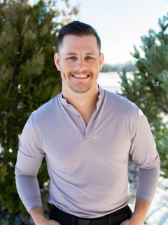 Man with hands in pockets smiling outdoors, wearing a gray shirt and black pants, snow in background.