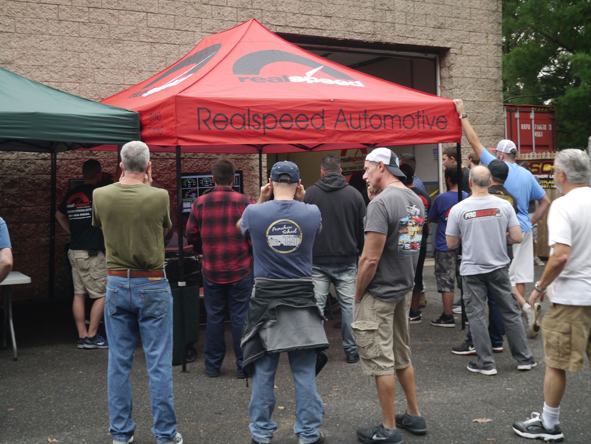 A group of men standing under a red canopy that says realspeed automotive
