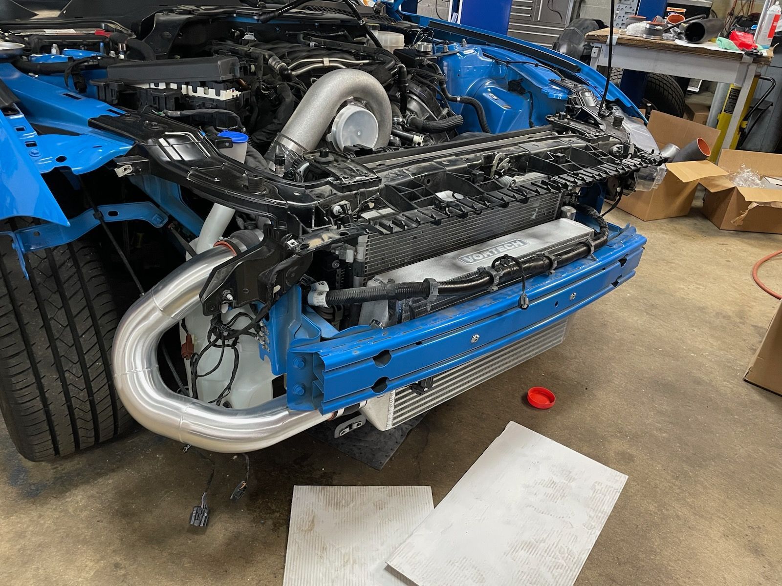 Blue car front with engine exposed, silver pipe, parts scattered, indoor setting.