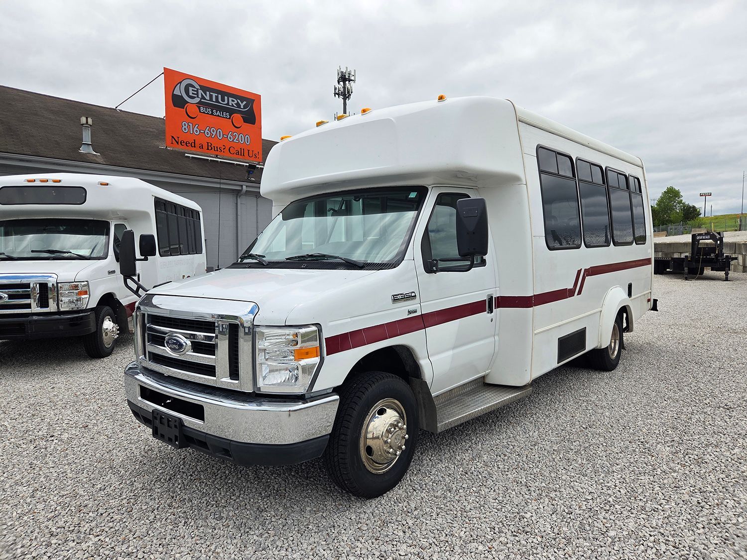 White shuttle bus with maroon stripe parked outside a dealership on a gravel lot