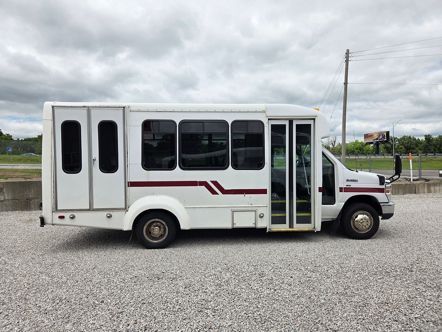 White shuttle bus parked on gravel under a cloudy sky, side doors open.