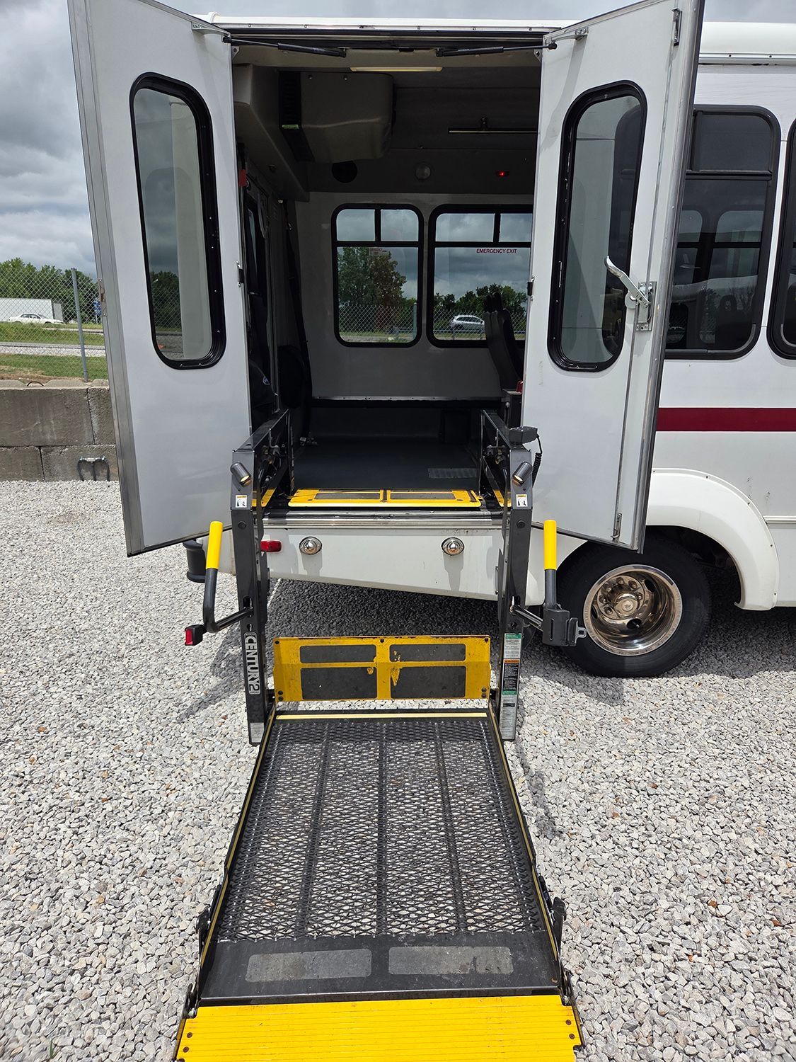 Wheelchair lift extended from a bus, with open rear doors on a gravel lot.