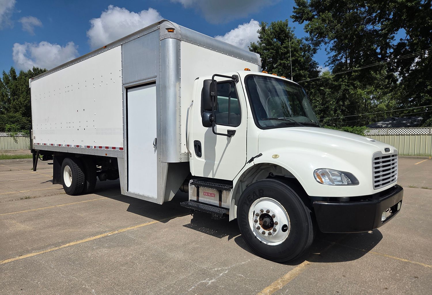 White box truck parked in a lot on a sunny day, side door open, with trees in the background