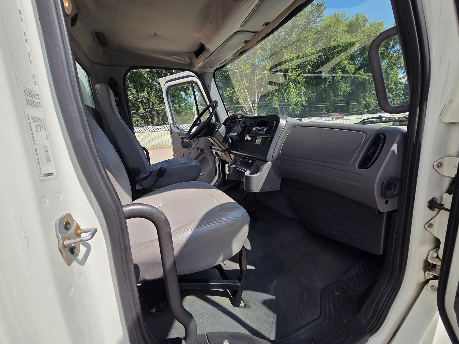 Inside a white vehicle, showing the driver’s seat, dashboard, and open side door with greenery outside.
