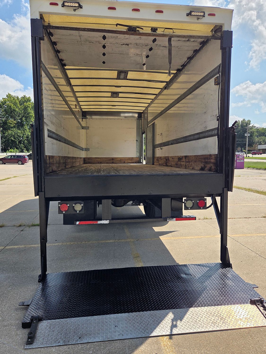 Open box truck with rear lift gate lowered on a sunny parking lot.