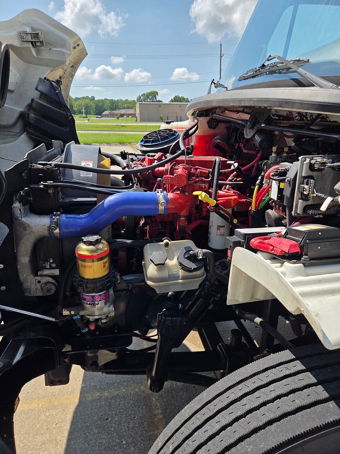 Open vehicle engine bay with red engine, blue hose, and visible front tire under daylight