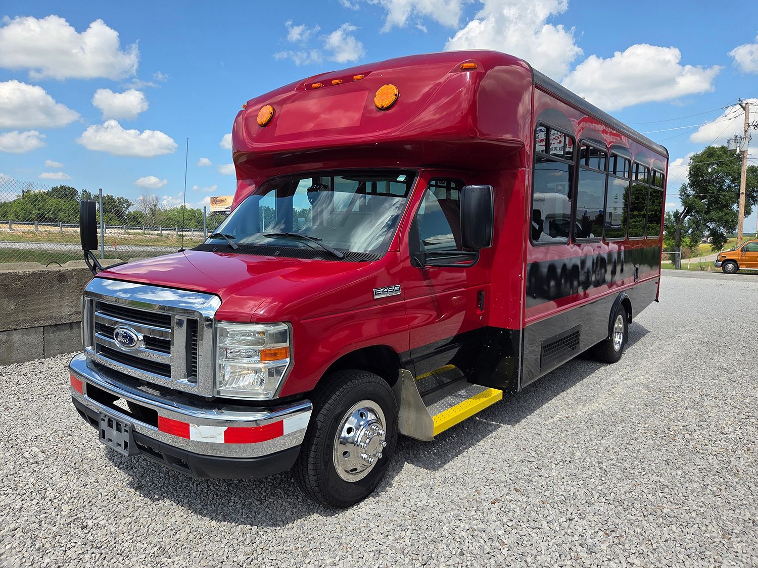 Red shuttle bus parked on gravel lot under a blue sky with clouds