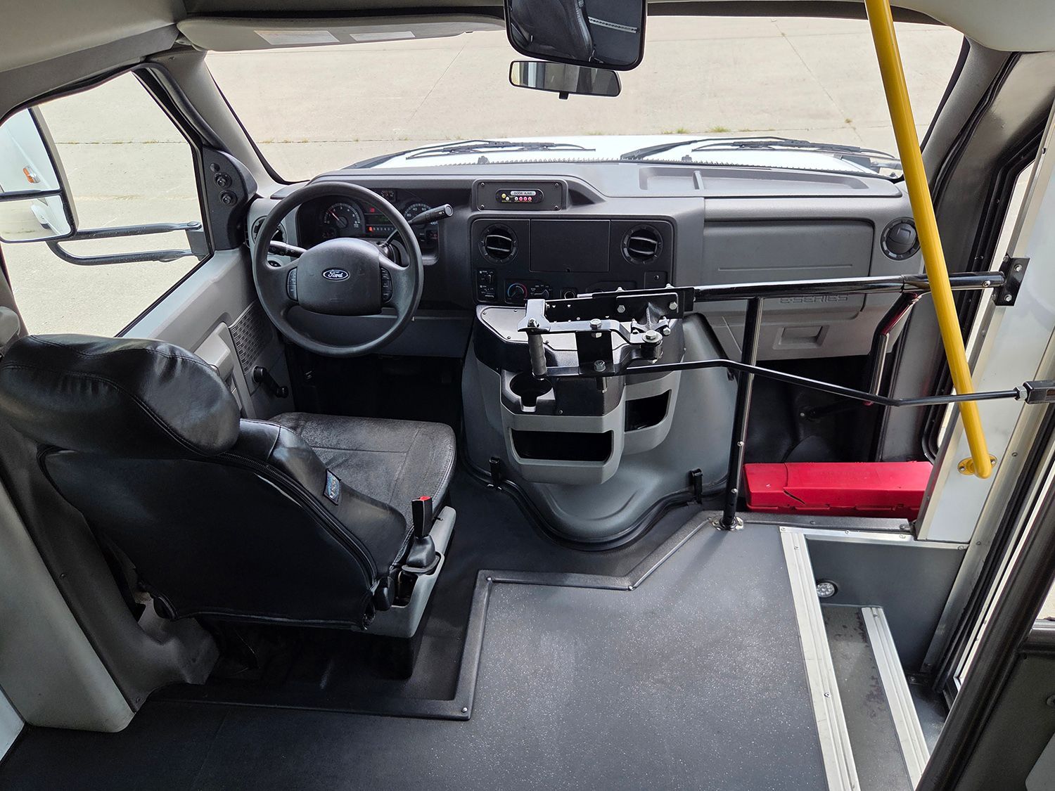Accessible bus interior with a wheelchair space, driver’s seat, steering wheel, and yellow support pole.