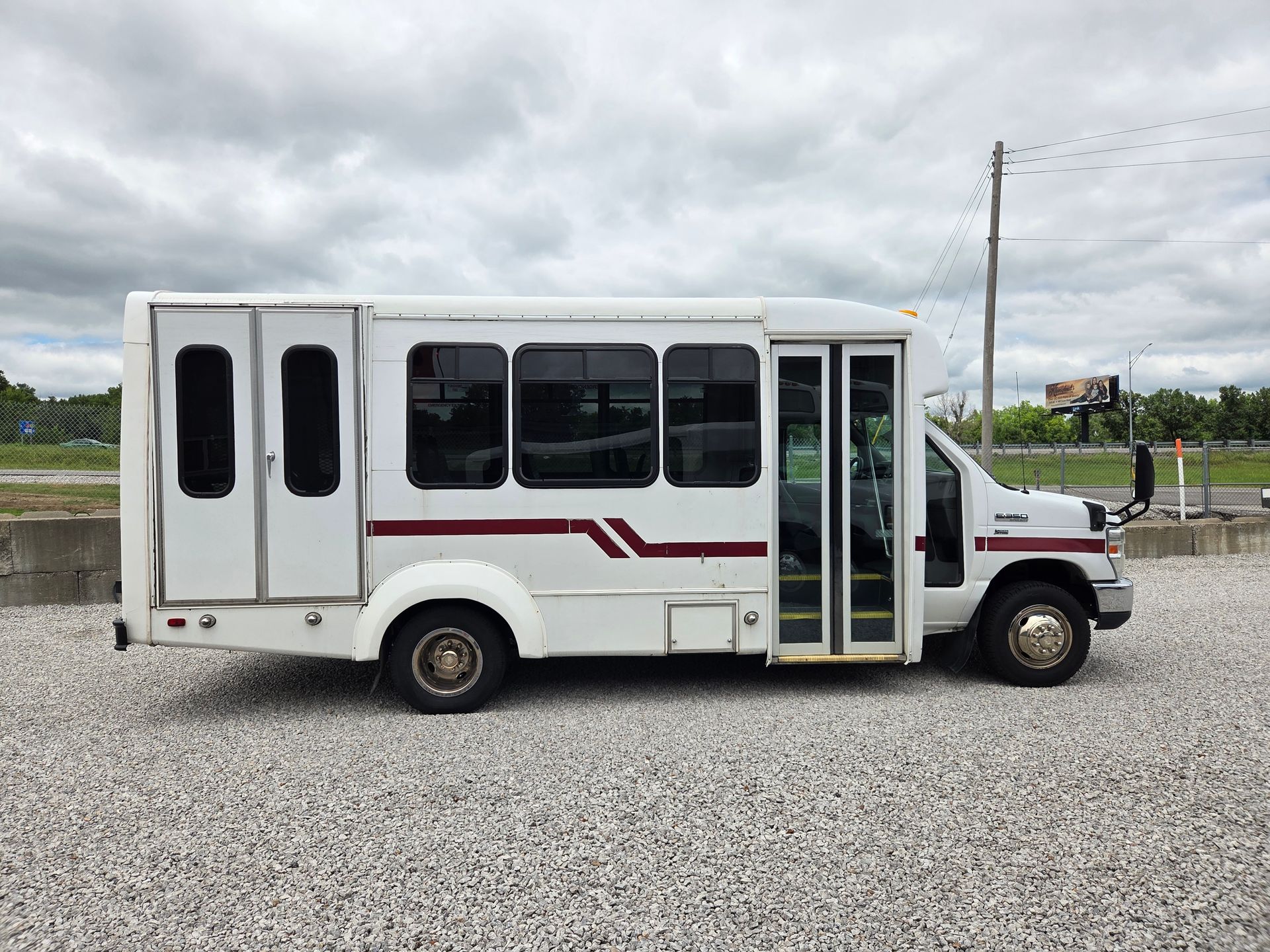 White shuttle bus with maroon stripes parked on gravel under cloudy sky, side view.