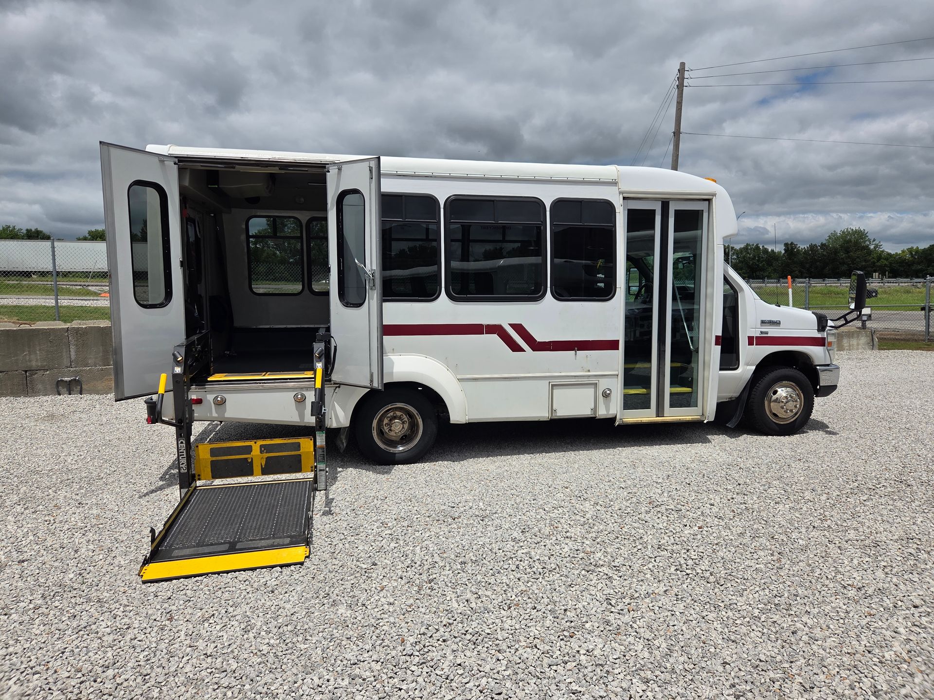 White shuttle bus with rear wheelchair lift ramp deployed on a gravel lot.