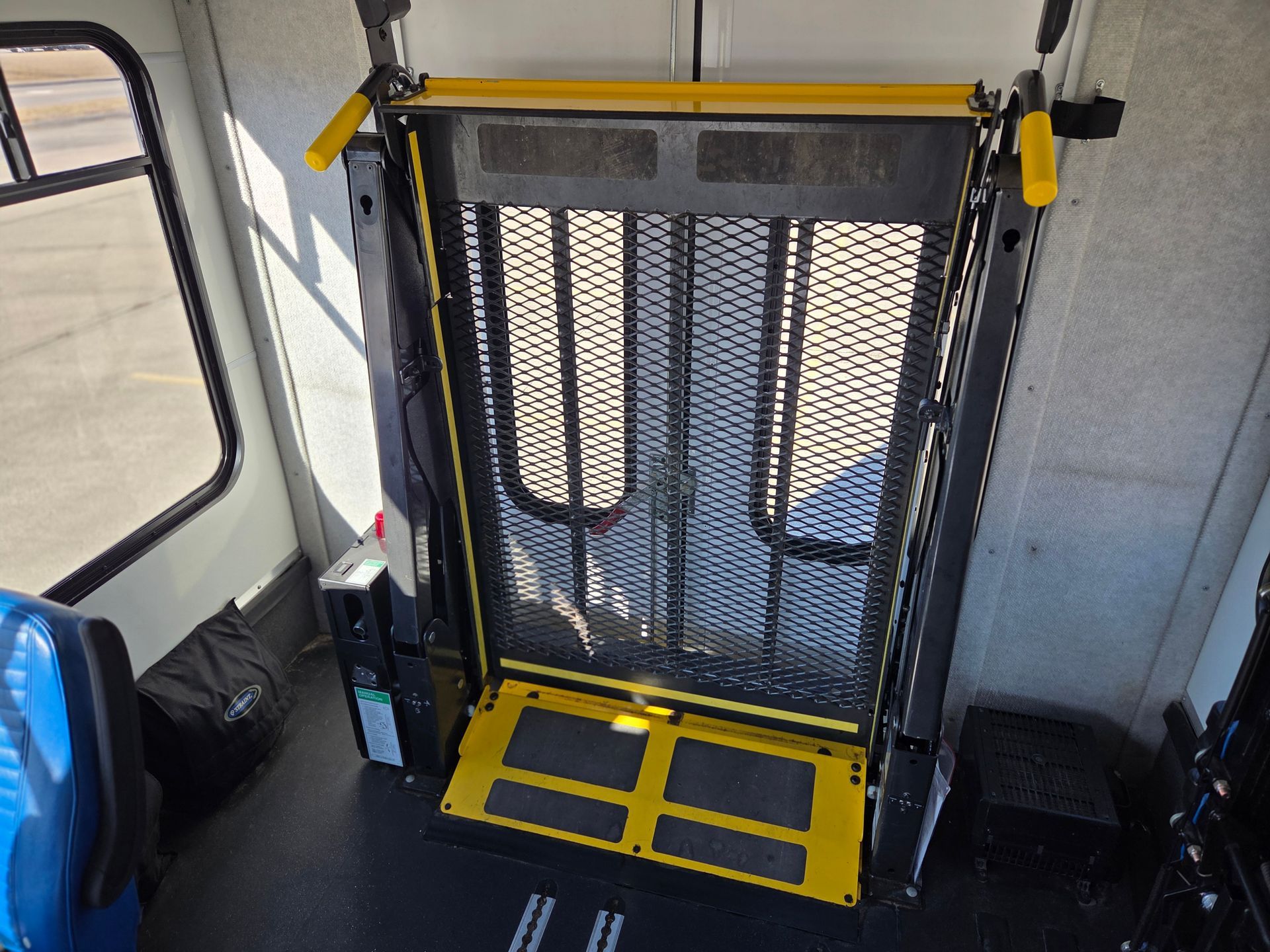 Fold-up wheelchair ramp with yellow safety frame inside a bus near the rear door.