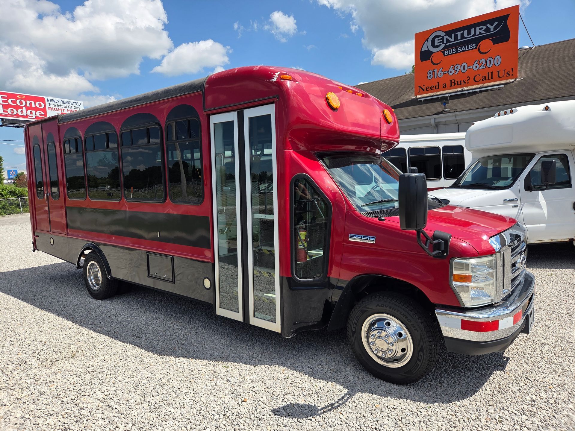 Maroon shuttle bus with black trim parked on a gravel lot under a blue sky