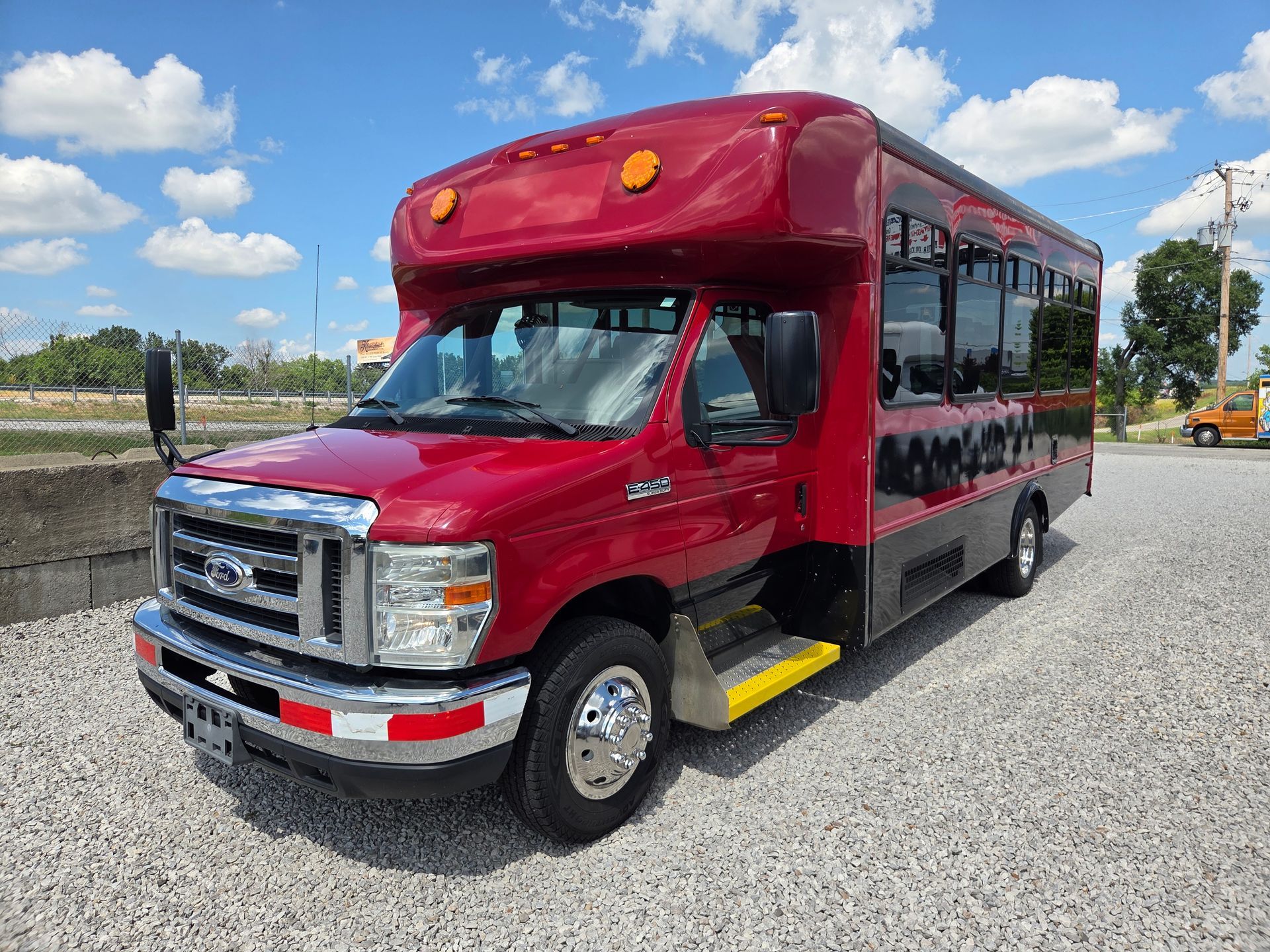 Red shuttle bus parked on gravel under a blue sky, with tinted windows and a chrome front grille.