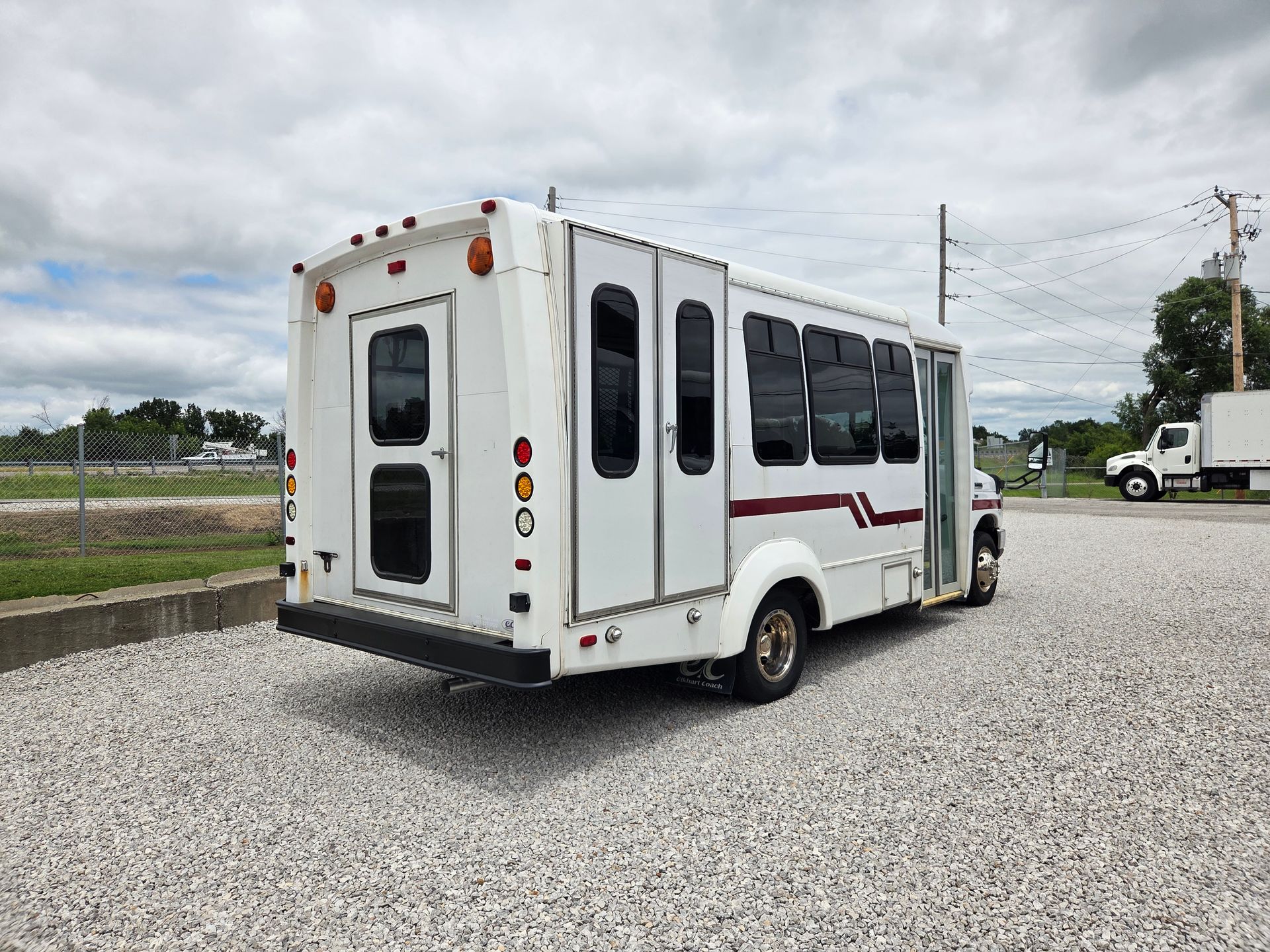 White shuttle bus with maroon stripe parked on gravel lot under cloudy sky