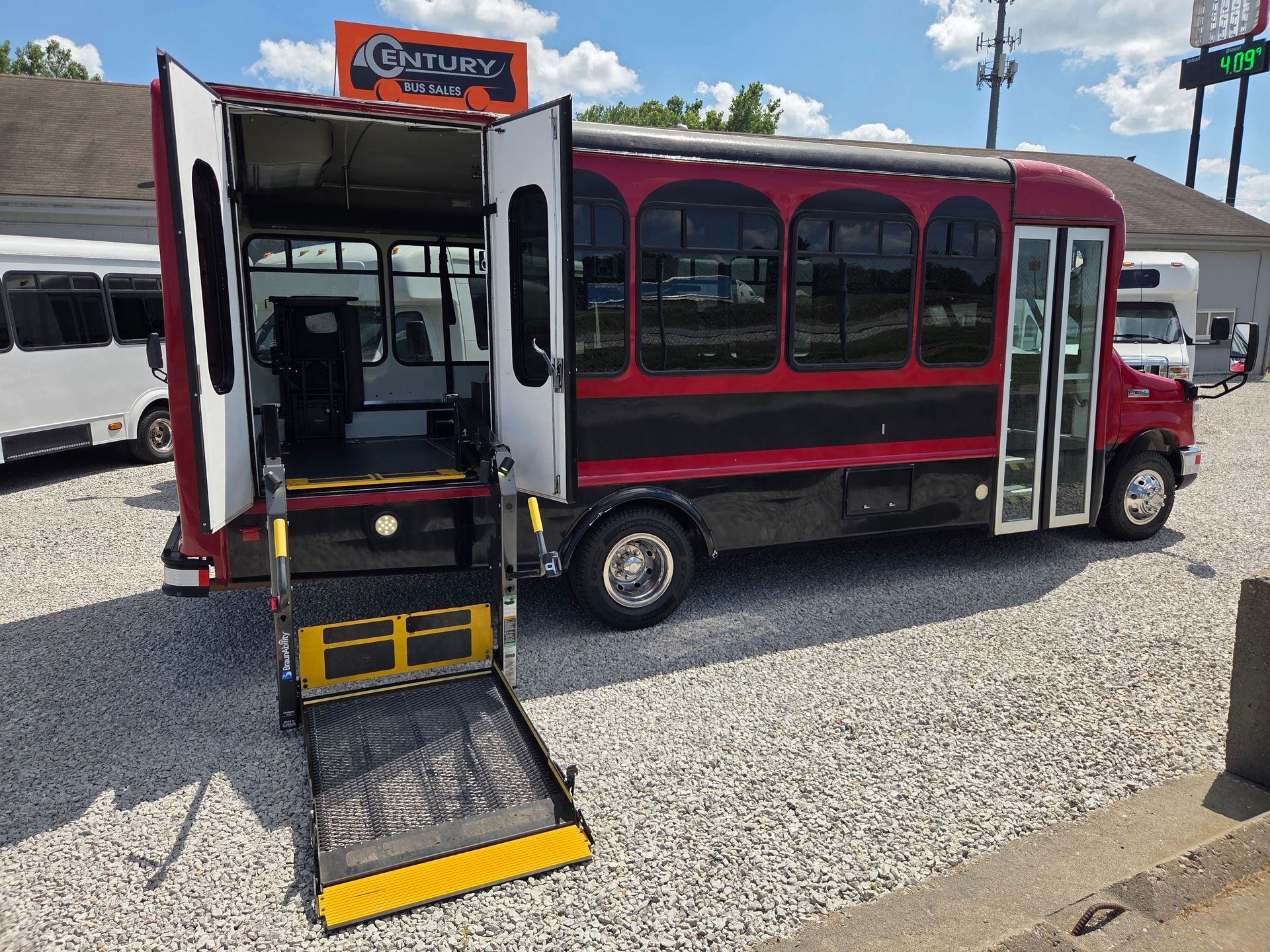 Red and black shuttle bus with wheelchair lift extended at a gravel lot.