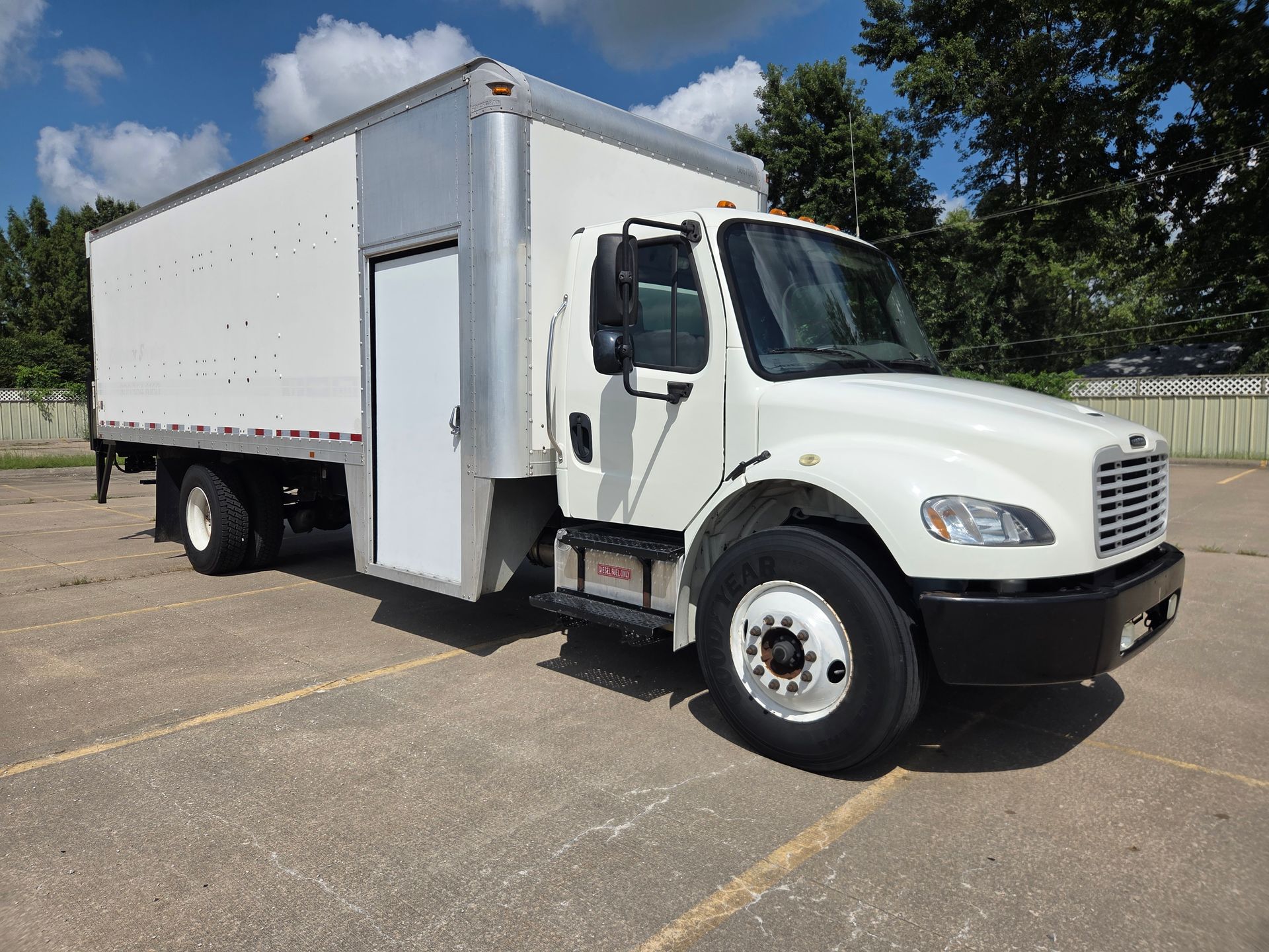 White box truck parked on a sunny lot, angled front view, with trees and blue sky in the background
