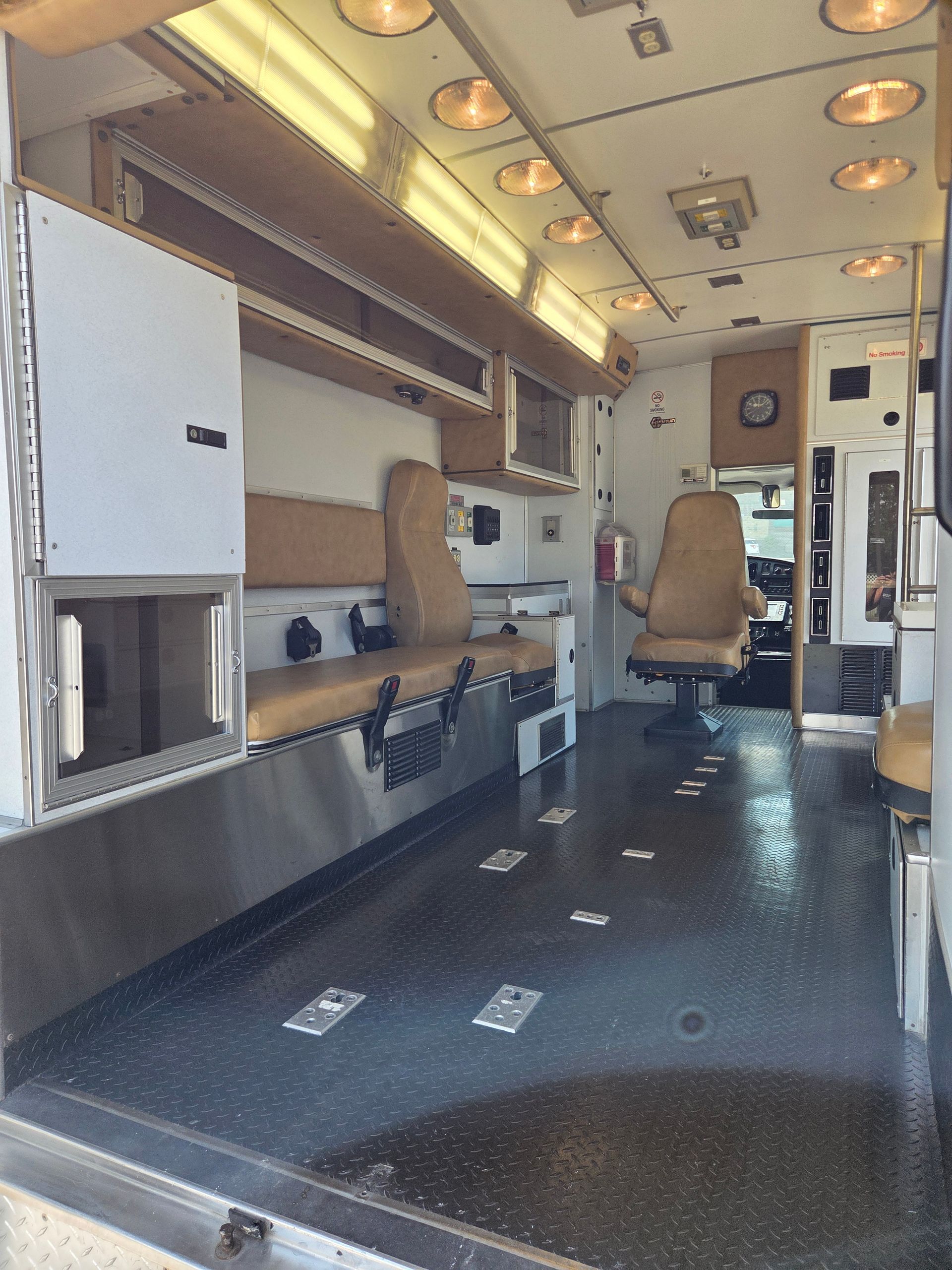 Interior of a small camper van with built-in cabinets, overhead lights, and two tan swivel seats.