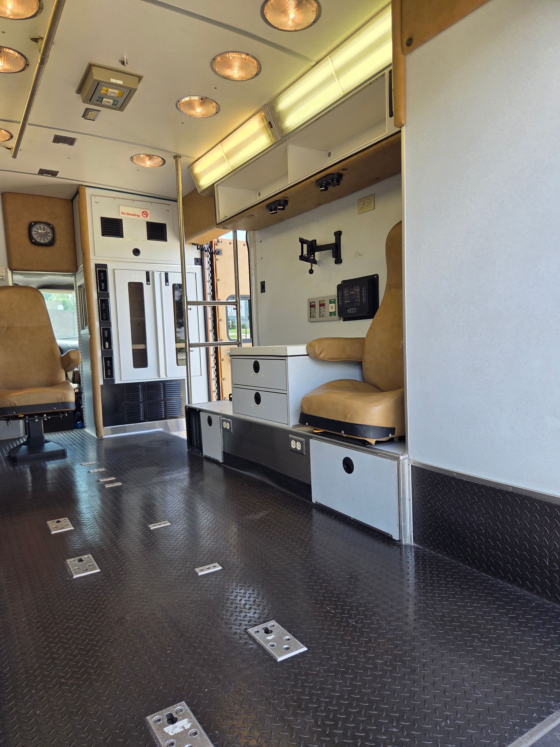 Inside an ambulance, showing stretchers, cabinets, and beige seats under bright overhead lights.