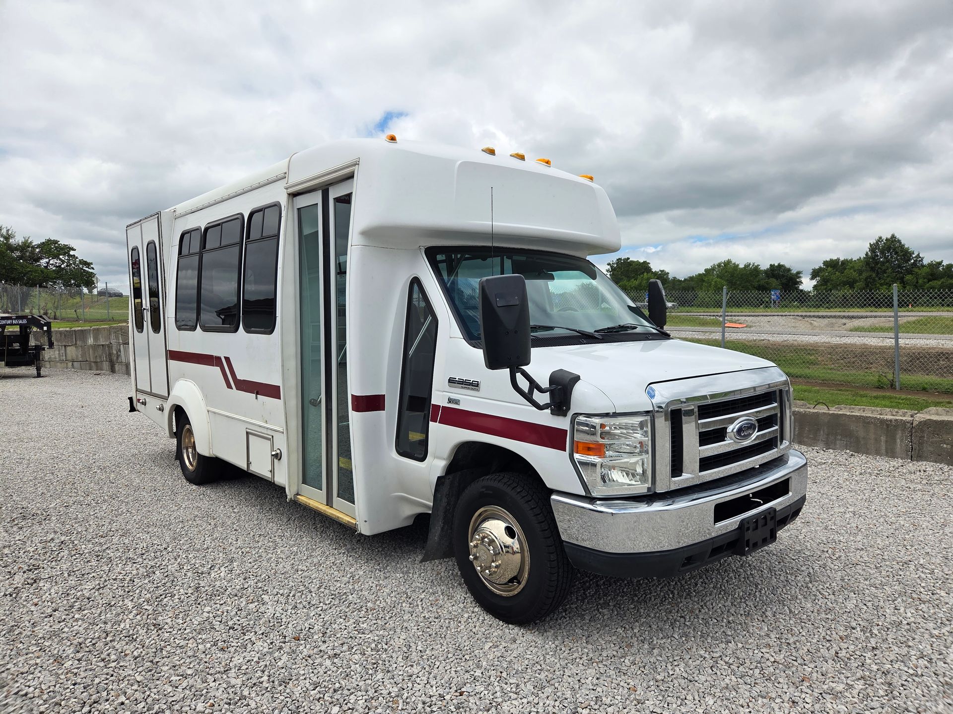 White shuttle bus parked on gravel under cloudy sky, with tinted windows and red stripe along the side