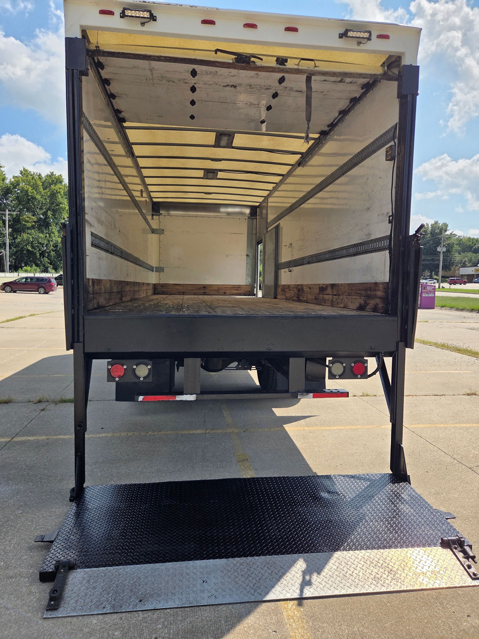 Open box truck with lowered lift gate, viewed from the rear in a parking lot.