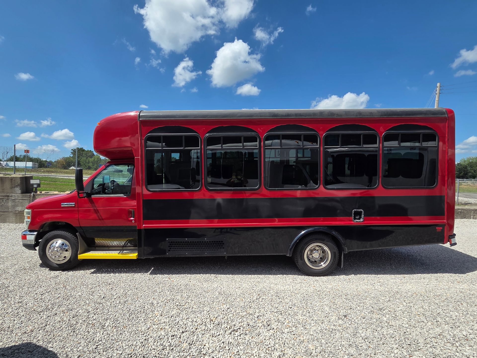 Red and black shuttle bus parked on a gravel lot under a blue sky.