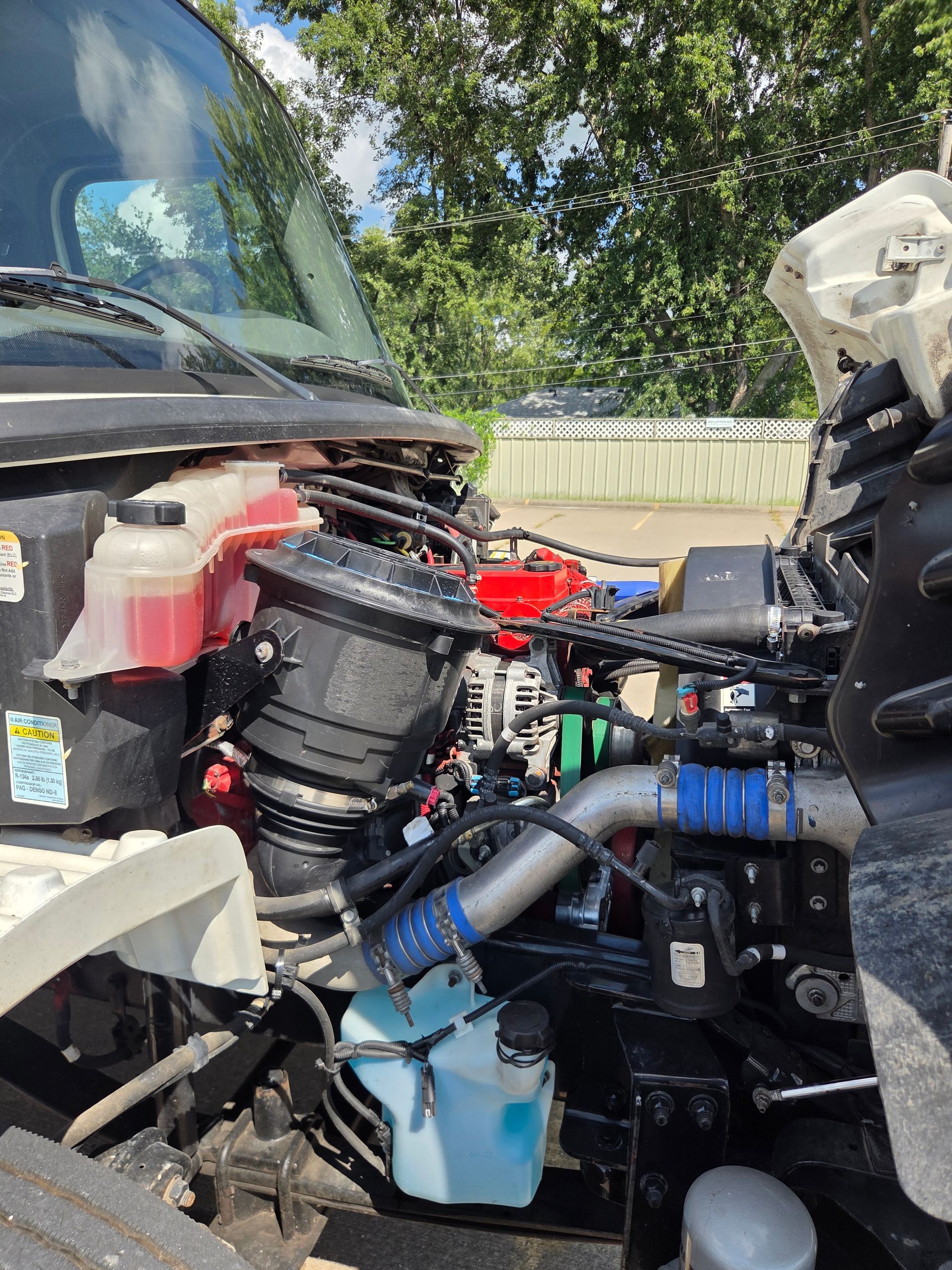 Open truck engine bay with exposed parts, blue coolant reservoir, and trees in the background.