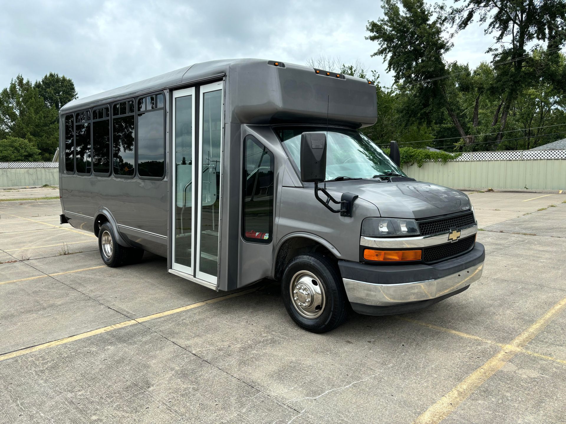 Silver shuttle bus parked in an empty lot, front door open, with trees in the background