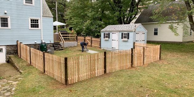 A person working in a backyard enclosed by a new wooden picket fence, next to a small blue shed and a house.