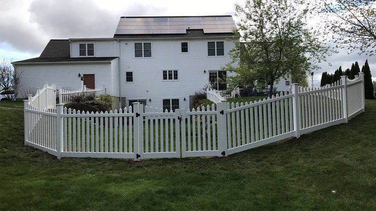 A white house with solar panels on the roof is enclosed by a white picket fence in a grassy yard under a cloudy sky.