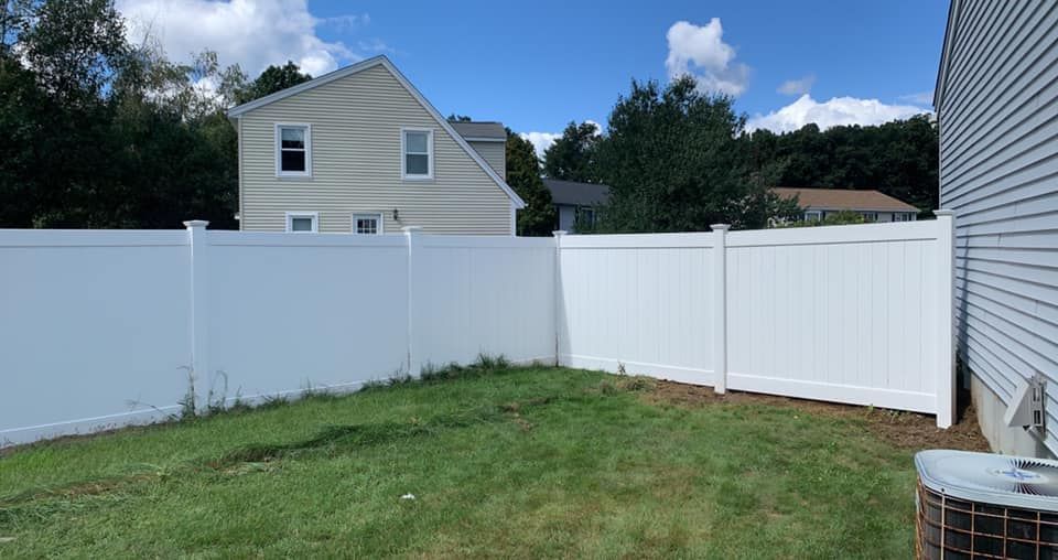 A white vinyl privacy fence encloses a green grassy backyard with a multi-story house visible in the background.