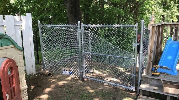 A chain-link fence gate installed outdoors next to a wooden deck with a blue plastic slide and a toy playhouse.