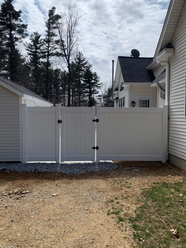 White vinyl double gate installed between two siding-clad houses, set against a background of trees and gravel.